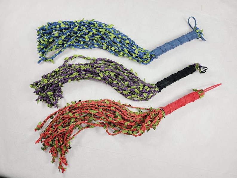 May include: Three decorative whips featuring long, braided strands in blue, purple, and red, each adorned with small green fabric leaves. The handles are wrapped in matching solid-colored fabric, and each whip includes a small loop at the end.