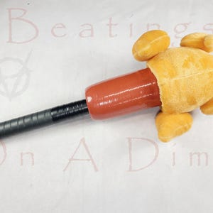 Può includere: Un oggetto di novità con una testa di pollo gialla di peluche attaccata a un corpo cilindrico rosso. Il corpo si trasforma in un manico nero e strutturato. Le parole "Beatings On A Dime" sono debolmente visibili sullo sfondo.