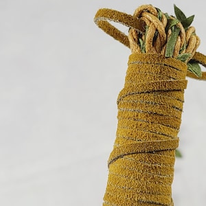 May include: A cylindrical handle wrapped in mustard yellow suede leather cord. The top features a decorative knot made from the same yellow cord, intertwined with thin green ribbon accents that extend outward in a circular loop pattern.