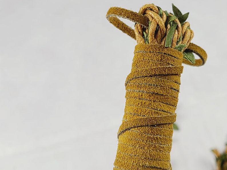 May include: A cylindrical handle wrapped in mustard yellow suede leather cord. The top features a decorative knot made from the same yellow cord, intertwined with thin green ribbon accents that extend outward in a circular loop pattern.