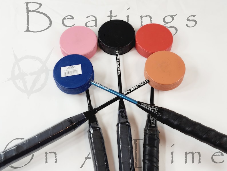 May include: A selection of badminton rackets and coloured pucks on a white background. The pucks are pink, black, red, blue, and orange. The rackets have black handles and blue shafts. The word "Beatings" is visible in the background.