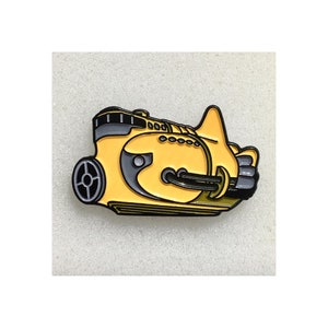 May include: A yellow and grey enamel pin shaped like a stylised submarine. The design features a prominent yellow hull with grey accents, a circular porthole, a small fin on top, and a rear propeller assembly, all outlined in bold black metal lines.