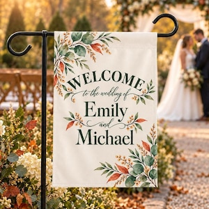 May include: A rectangular off-white garden flag hanging on a black metal stand. The flag features dark green text reading "WELCOME to the wedding of Emily and Michael" surrounded by watercolour illustrations of green leaves and orange berries.