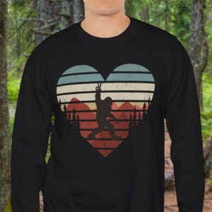 May include: Black sweatshirt with a vintage-style heart design featuring a silhouette of Bigfoot making a peace sign in front of a mountain range. The heart is filled with red, blue, and white stripes.
