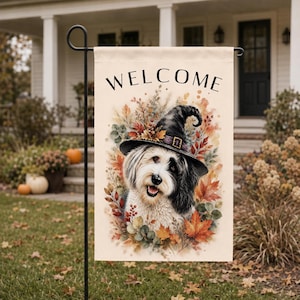 May include: A rectangular garden flag featuring the word WELCOME in black text at the top. The center displays a fluffy black and white dog wearing a pointed witch hat, surrounded by a watercolor illustration of colorful autumn leaves and berries.