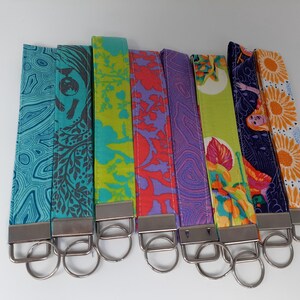 May include: Eight colorful fabric keychains with silver key rings. The keychains feature different patterns, including floral, geometric, and abstract designs.