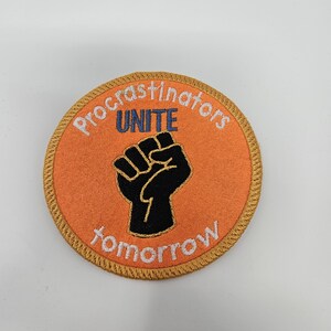 May include: A round orange patch with a gold border. The patch has a black fist raised in the air with the words "Procrastinators Unite tomorrow" around it.