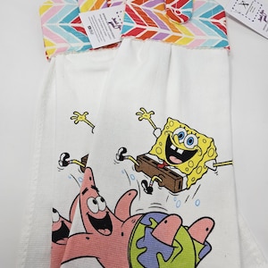 May include: Two white kitchen towels with a colorful chevron pattern on the top. The towels feature a printed image of Spongebob Squarepants and Patrick Star from the cartoon series Spongebob Squarepants.
