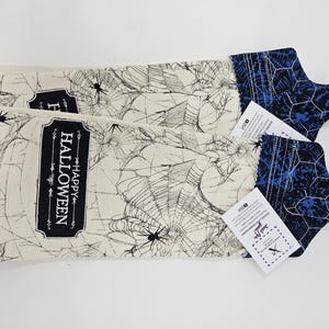 May include: Two Halloween-themed kitchen towels. The towels feature a spiderweb design with spiders and the words "HAPPY HALLOWEEN" in a black frame. The ends are blue with a geometric pattern.