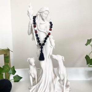 May include: A white statue of a woman holding a torch, with two dog statues at her feet. A black beaded necklace with red accents and a black tassel hangs around the statue's neck. The statue is on a white shelf.