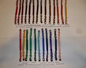 Op de afbeelding: Een kleurenkaart met lederen armbandmonsters in verschillende kleuren, waaronder oxblood, maroon, mocassinkleur, chocolade, roestbruin, bruin, buckskin, geel, groen, turquoise, marineblauw, zwart en koraalroze. Het leer is gevlochten en heeft zilveren metalen sluitingen.