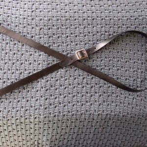 May include: A black leather guitar strap with a gold metal buckle. The strap is laid out in a criss-cross pattern on a gray textured surface.
