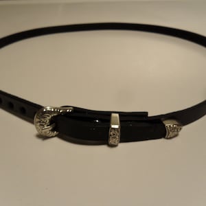May include: A black leather belt with a silver-colored buckle and accents. The belt has a classic design with a decorative buckle and two matching silver accents. The belt is approximately 0.75 inches wide.