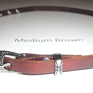May include: A brown leather belt with silver heart-shaped studs and a silver buckle. The buckle has a decorative design. The belt is labeled "Medium Brown".