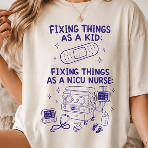 May include: A cream-colored t-shirt featuring blue text and graphics. The text reads: FIXING THINGS AS A KID: followed by an adhesive bandage illustration, and FIXING THINGS AS A NICU NURSE: with illustrations of an incubator, monitor, stethoscope, pacifier, baby bottle, and footprints.