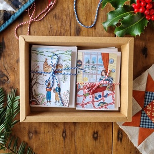 May include: A set of illustrated greeting cards in a brown box. The cards are tied with blue and red twine. One card shows a winter scene with people, and the other shows a person by a window. Holly and a quilt are in the background.