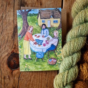 Könnte beinhalten: Grußkarte mit einer Illustration von drei Personen, die im Freien stricken, mit Garn und Strickzubehör auf einem Tisch. Eine Strähne grünes Garn befindet sich im Vordergrund. Die Szene spielt in einem Garten mit einem Cottage im Hintergrund.
