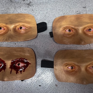 May include: Four prosthetic eye masks with a flesh-toned base. Two masks have realistic red-veined eyes, while the other two feature open wounds with dripping blood. Each mask has a black elastic strap for wear.
