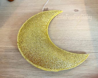 Op de afbeelding: Een gouden glitter halve maanvormige ornament. De maan is gemaakt van vilt en heeft een touwtje aan de bovenkant om op te hangen.