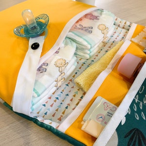 May include: A yellow changing mat with multiple pockets for baby supplies. The mat has a white trim and a green and white patterned fabric lining. The pockets contain nappies, a dummy, a washcloth, and a tube of baby cream.
