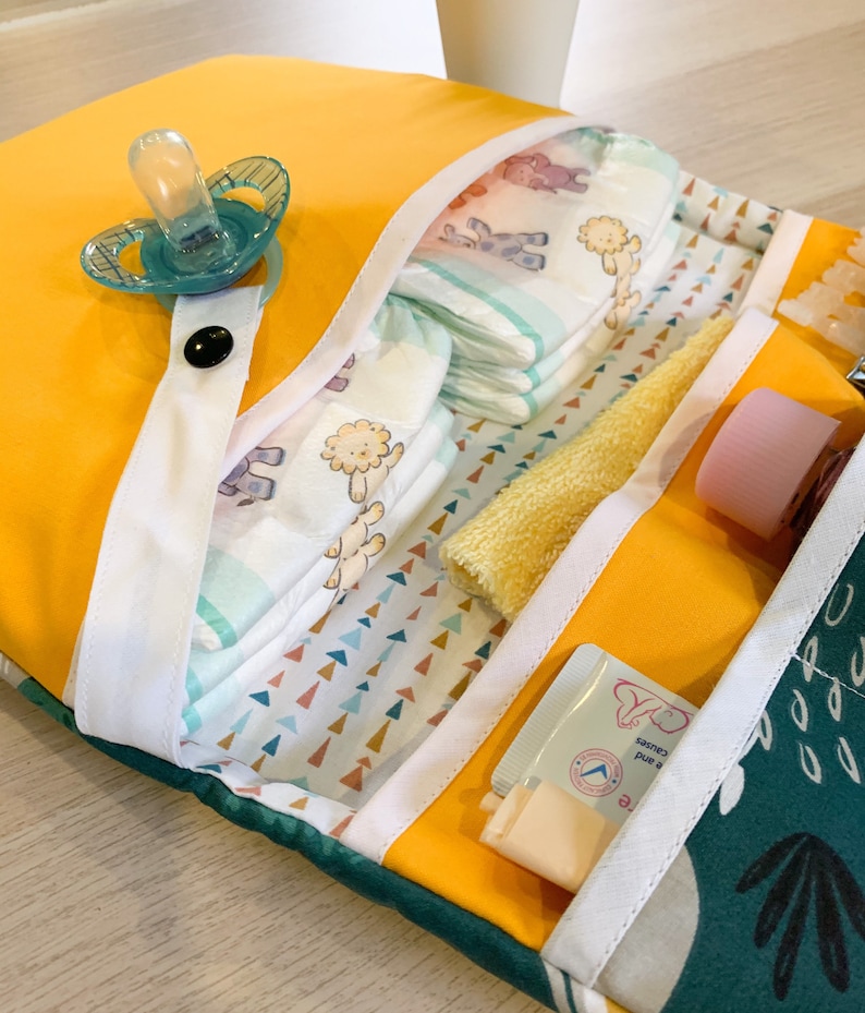 May include: A yellow changing mat with multiple pockets for baby supplies. The mat has a white trim and a green and white patterned fabric lining. The pockets contain nappies, a dummy, a washcloth, and a tube of baby cream.