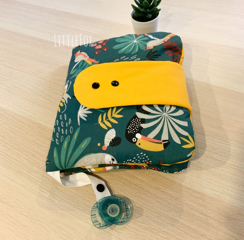 May include: A green and yellow fabric nappy changing mat with a toucan, leaves, and flowers print. The mat has a yellow flap with two black buttons. The mat is folded and has a white strap with a black snap and a blue dummy attached.