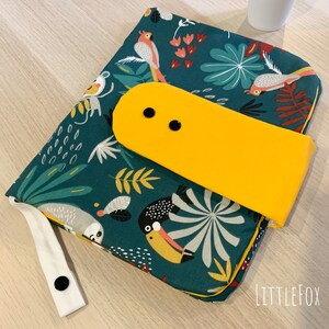 May include: A green changing mat with a tropical print featuring toucans, sloths, and parrots. The mat has a yellow flap with two black buttons and a white strap with a black button.
