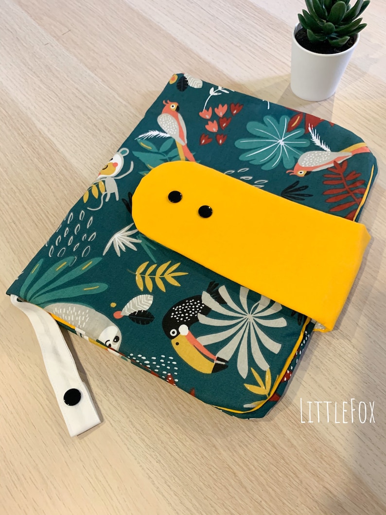 May include: A green changing mat with a tropical print featuring toucans, sloths, and parrots. The mat has a yellow flap with two black buttons and a white strap with a black button.