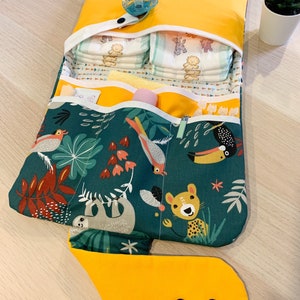 May include: A yellow changing mat with a jungle animal print. The mat has multiple pockets and a flap that folds over to create a changing station. The mat is open and shows a stack of nappies, a dummy, and other baby supplies.
