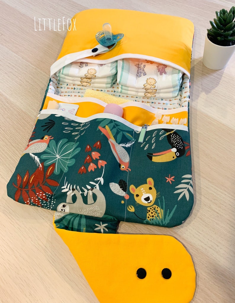May include: A yellow changing mat with a jungle animal print. The mat has multiple pockets and a flap that folds over to create a changing station. The mat is open and shows a stack of nappies, a dummy, and other baby supplies.