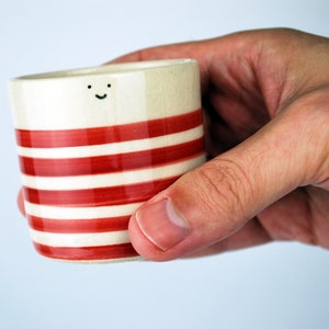 May include: A small, ceramic cup with a cream-colored base and horizontal red stripes. The cup has a small, smiling face near the top. The cup is held in a hand against a light blue background. The cup is approximately 3 inches tall.