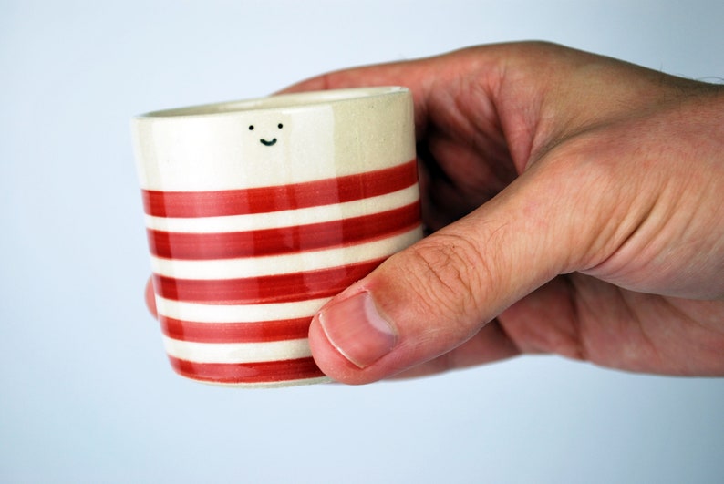 May include: A small, ceramic cup with a cream-colored base and horizontal red stripes. The cup has a small, smiling face near the top. The cup is held in a hand against a light blue background. The cup is approximately 3 inches tall.