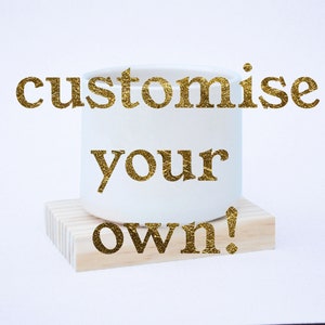 May include: White ceramic planter with the words "customize your own!" in gold lettering. The planter is sitting on a wooden base.