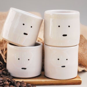 May include: Four off-white ceramic cups with minimalist faces, each featuring two black dots for eyes and a black dash for a mouth. The cups are stacked on a wooden surface, with coffee beans scattered around.