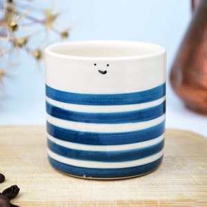 Könnte beinhalten: Eine Keramik-Kaffeetasse mit weißem Grund und blauen horizontalen Streifen. Die Tasse hat ein kleines, lächelndes Gesicht aufgemalt. Die Tasse steht auf einem Holzschneidebrett mit Kaffeebohnen.
