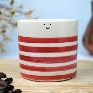 May include: A ceramic coffee mug with a cream-colored base and horizontal red stripes. The mug has a small, smiling face drawn near the top. Coffee beans are scattered on a wooden surface in the foreground.