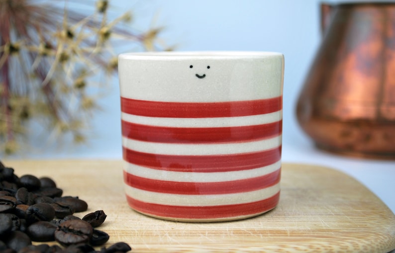 May include: A ceramic coffee mug with a cream-colored base and horizontal red stripes. The mug has a small, smiling face drawn near the top. Coffee beans are scattered on a wooden surface in the foreground.