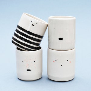 May include: Four white ceramic mugs with hand-painted faces. One mug has black stripes, one has red dots, one has a black mouth, and one has a black mouth and eyes.