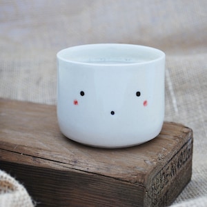 May include: A white ceramic mug with a simple hand-painted face. The mug has two black eyes, a small black mouth, and two small red cheeks.