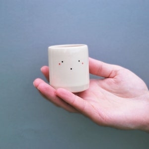 May include: A small, white ceramic cup with a simple, hand-painted face. The cup has two small red dots for cheeks and two black dots for eyes.