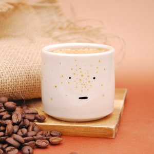 May include: A white ceramic coffee mug with a speckled brown glaze and a hand-painted face. The mug is filled with coffee and is sitting on a wooden coaster.