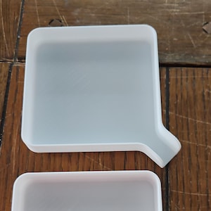 May include: Two light gray, square-shaped trays with a white border and a spout on one side. The trays are made of a translucent material and are placed on a wooden surface.