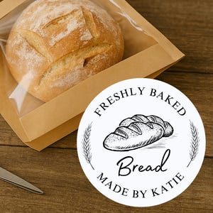 May include: A round loaf of freshly baked bread in a brown paper bag with a clear window. A white sticker with the text "FRESHLY BAKED Bread MADE BY KATIE" is visible. A pair of scissors is on the wooden surface.
