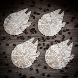 Peut inclure: Quatre sous-verres en bois blanc avec un contour détaillé du vaisseau spatial Millennium Falcon de Star Wars. Les sous-verres sont entourés de grains de café sur une surface en bois brun.