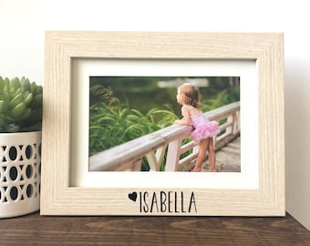 May include: A light wood picture frame with a white mat and a photo of a young girl in a pink dress standing on a wooden bridge. The frame has a black heart and the name "ISABELLA" written below the photo.