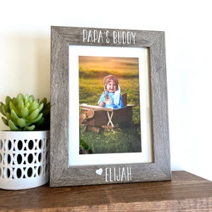 May include: A gray wooden picture frame with a white inscription that reads "Papa's Buddy" and a heart symbol followed by the name "Elijah". The frame contains a photo of a baby wearing a blue shirt and aviator goggles sitting in a toy airplane.
