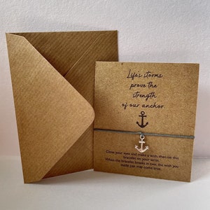 Peut inclure: Une carte brune avec le texte "Life's storms prove the strength of our anchor." Un charme d'ancre argenté est attaché à un bracelet en corde grise. La carte comprend également des instructions pour faire un vœu et attacher le bracelet à votre poignet.