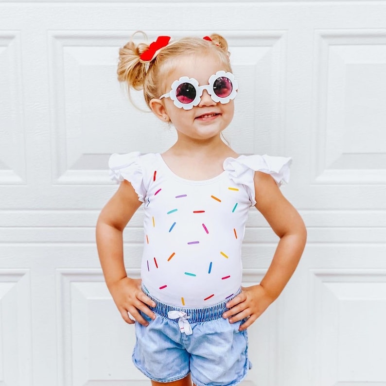 May include: A young girl wearing a white leotard with a colorful sprinkle pattern, denim shorts, and white flower-shaped sunglasses with pink lenses. She has red hair bows in her hair.