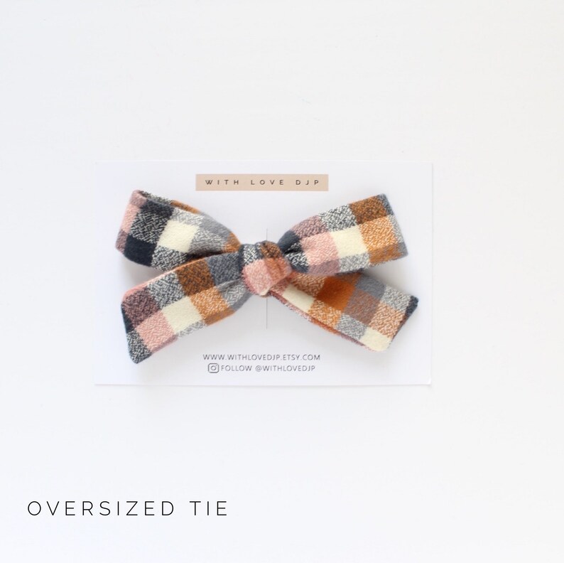 May include: Oversized tie hair bow in a plaid pattern with pink, orange, white, and gray colors. The bow is tied with a knot in the center. The text "WITH LOVE DJP" is printed on a white card behind the bow.
