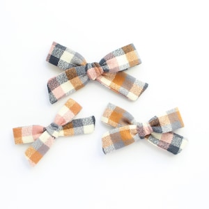 May include: Three plaid hair bows in different sizes. The bows are made of a soft, woven fabric with a pink, brown, and gray plaid pattern.